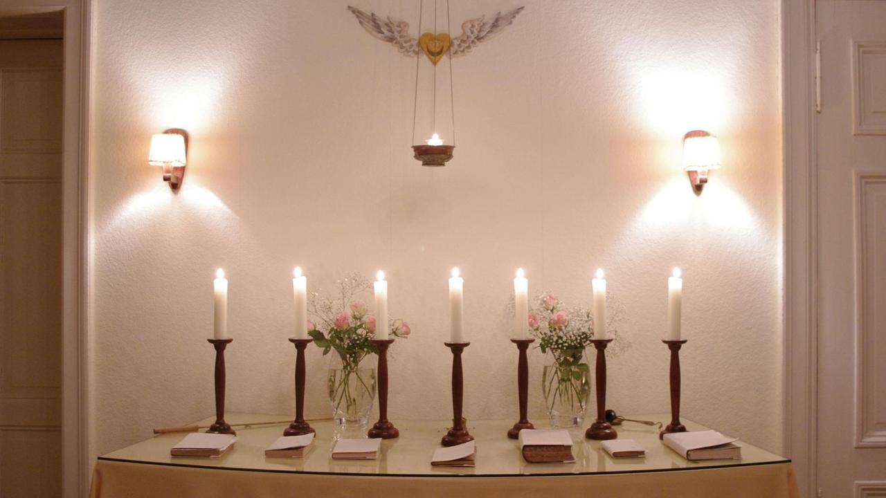 Auf dem Altar, einem schlichten Tisch mit hellem Tuch und einer Glasplatte, stehen sieben Leuchter mit brennenden Kerzen, vor jedem davon liegt ein Buch: die heilige Schrift der jeweils durch die Kerze repräsentierten Religion. Auf dem Altar, einem schlichten Tisch mit hellem Tuch und einer Glasplatte, stehen sieben Leuchter mit brennenden Kerzen, vor jedem davon liegt ein Buch: die heilige Schrift der jeweils durch die Kerze repräsentierten Religion.