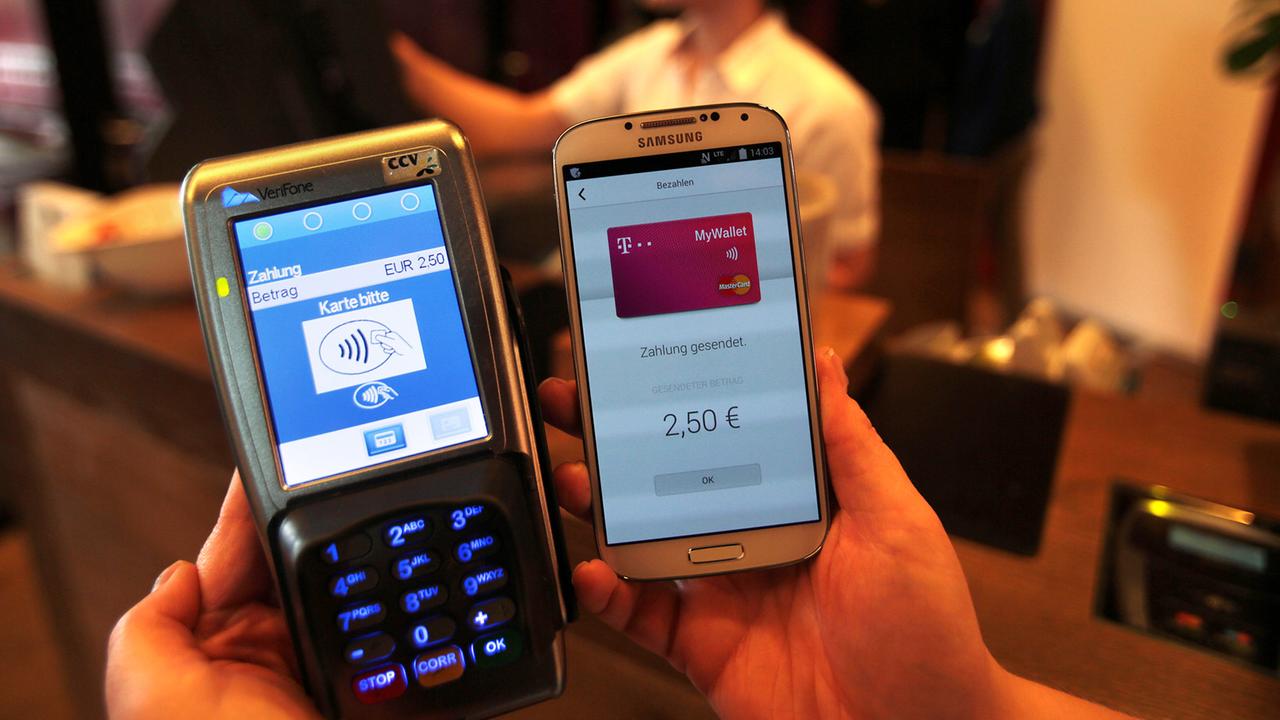 Eine Frau zeigt ein Smartphone mit der App MyWallet neben einem Empfangsgerät an der Kasse eines Restaurants. Eine Frau zeigt ein Smartphone mit der App MyWallet neben einem Empfangsgerät an der Kasse eines Restaurants.