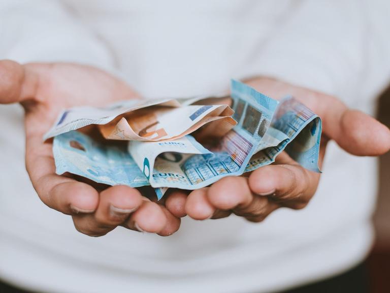 Eine Person im weißen Pullover hält die Hände zu einer nach oben geöffneten Schale. Darin liegen mehrere 20-Euro-Scheine und ein 50-Euro-Schein.