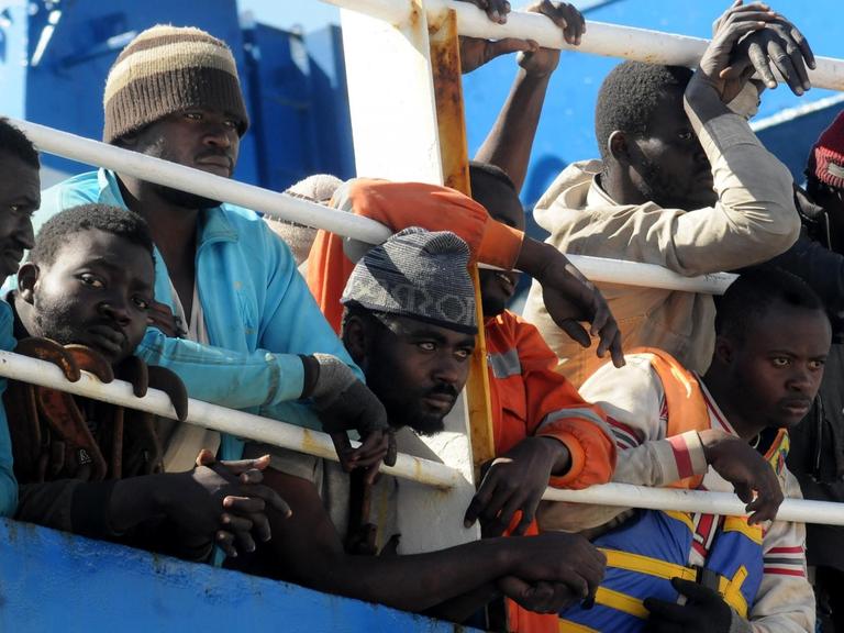 Flüchtlinge schauen auf einem Schiff durch die Stangen der Reeling