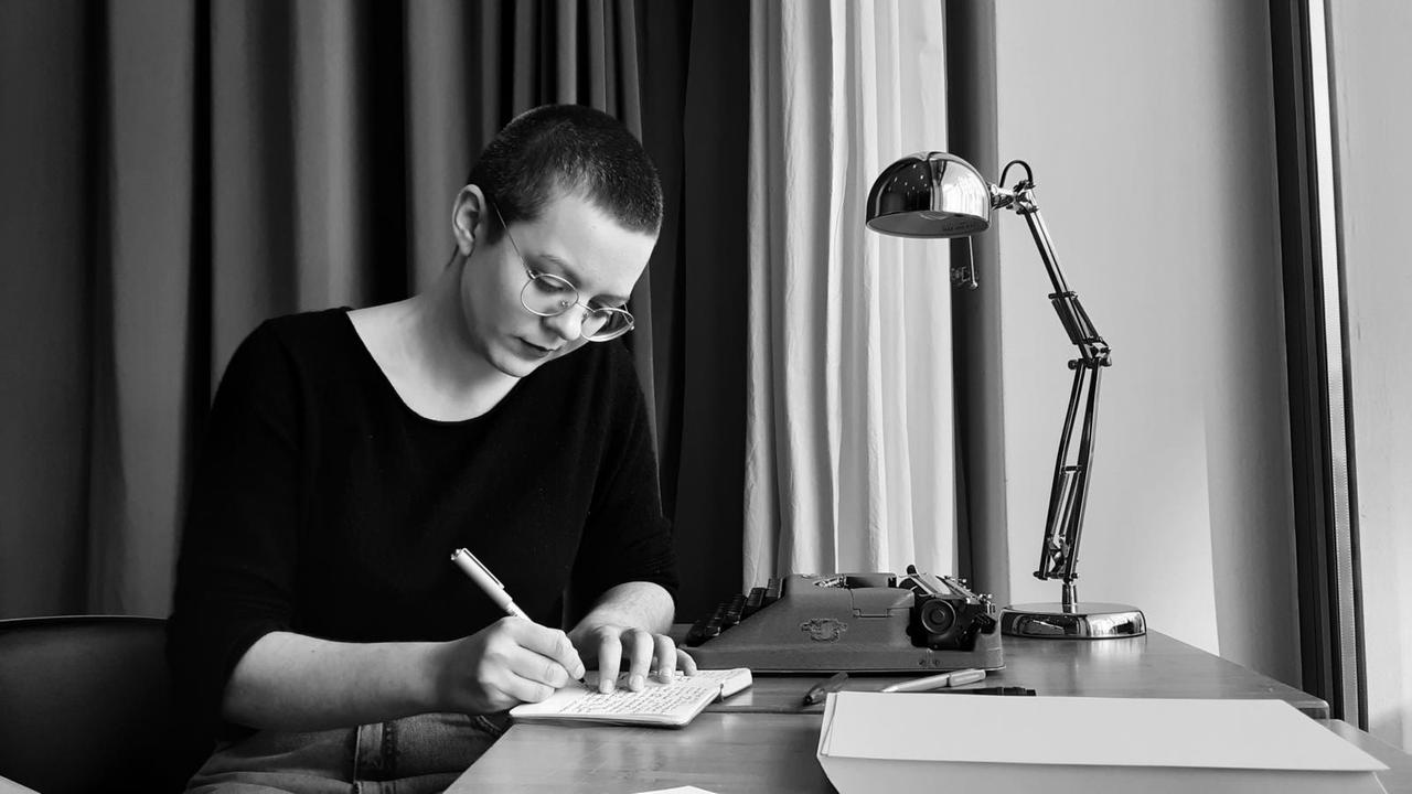 Eine junge Frau sitzt am Tisch vor einer Schreibmaschine und macht sich handschriftlich Notizen.