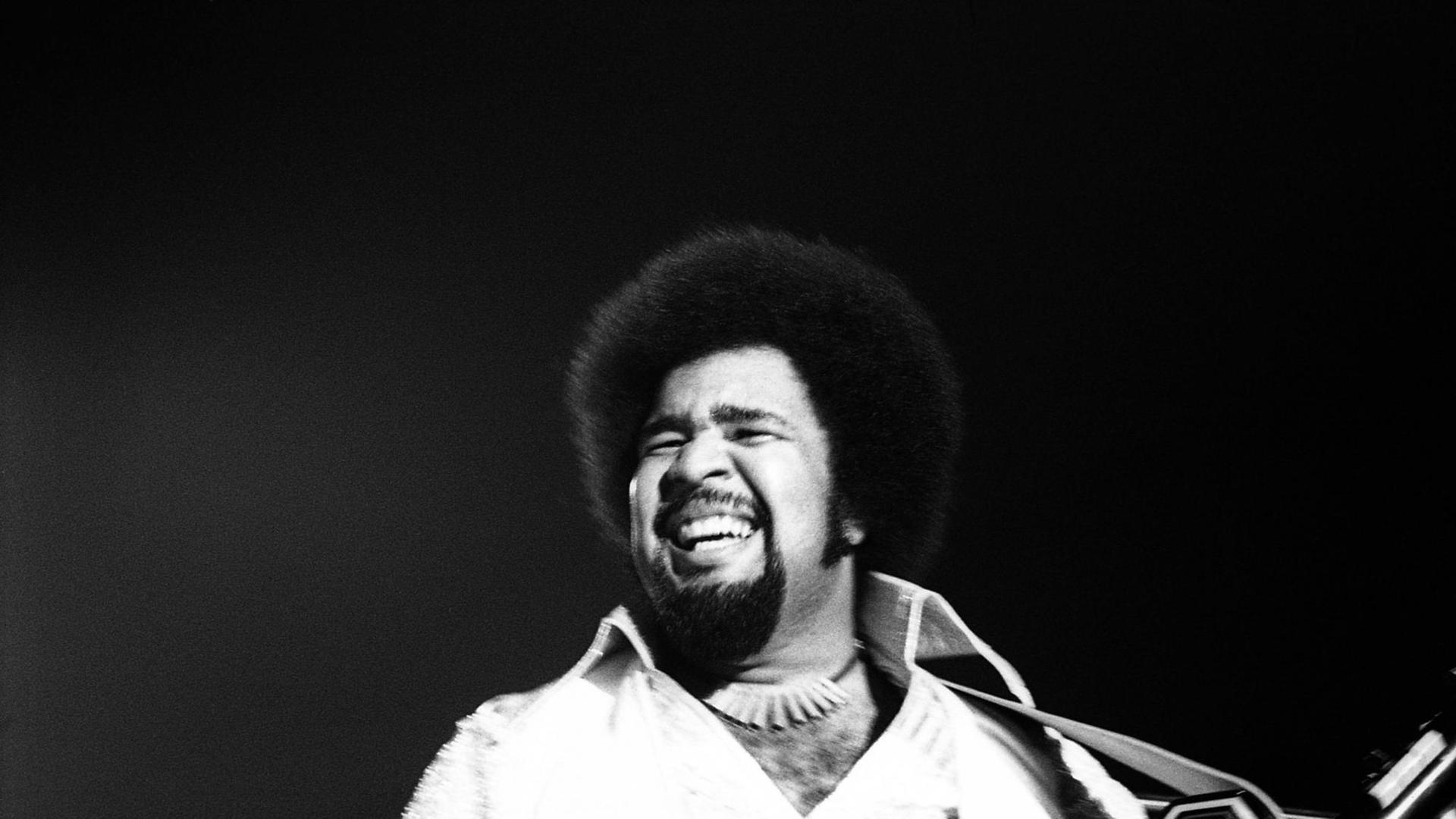 Das s/w-Bild zeigt einen Mann mit Vollbart und Afro, der ein Umhängekeyboard hält. Er blickt lachend in die Ferne. Das s/w-Bild zeigt einen Mann mit Vollbart und Afro, der ein Umhängekeyboard hält. Er blickt lachend in die Ferne.