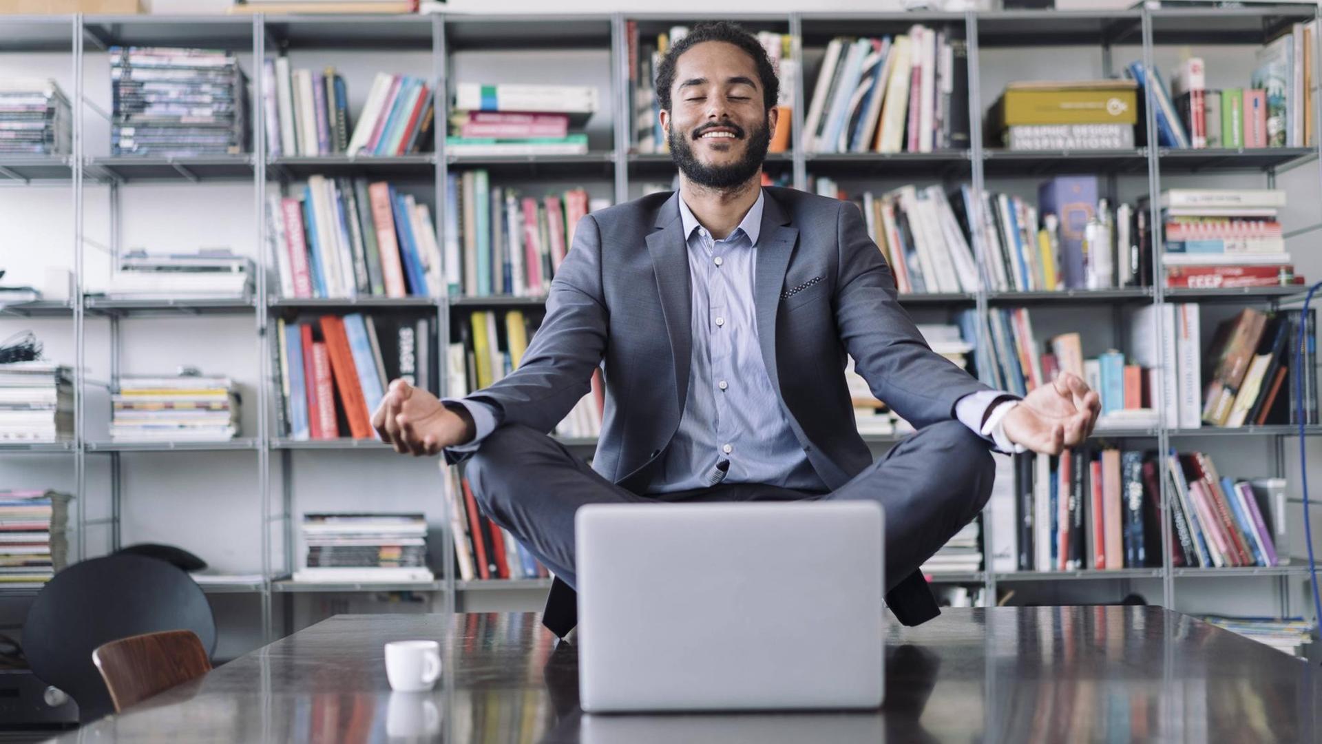 Ein junger Geschäftsmann sitzt in Meditationshaltung auf seinem Arbeitstisch hinter seinem Laptop.