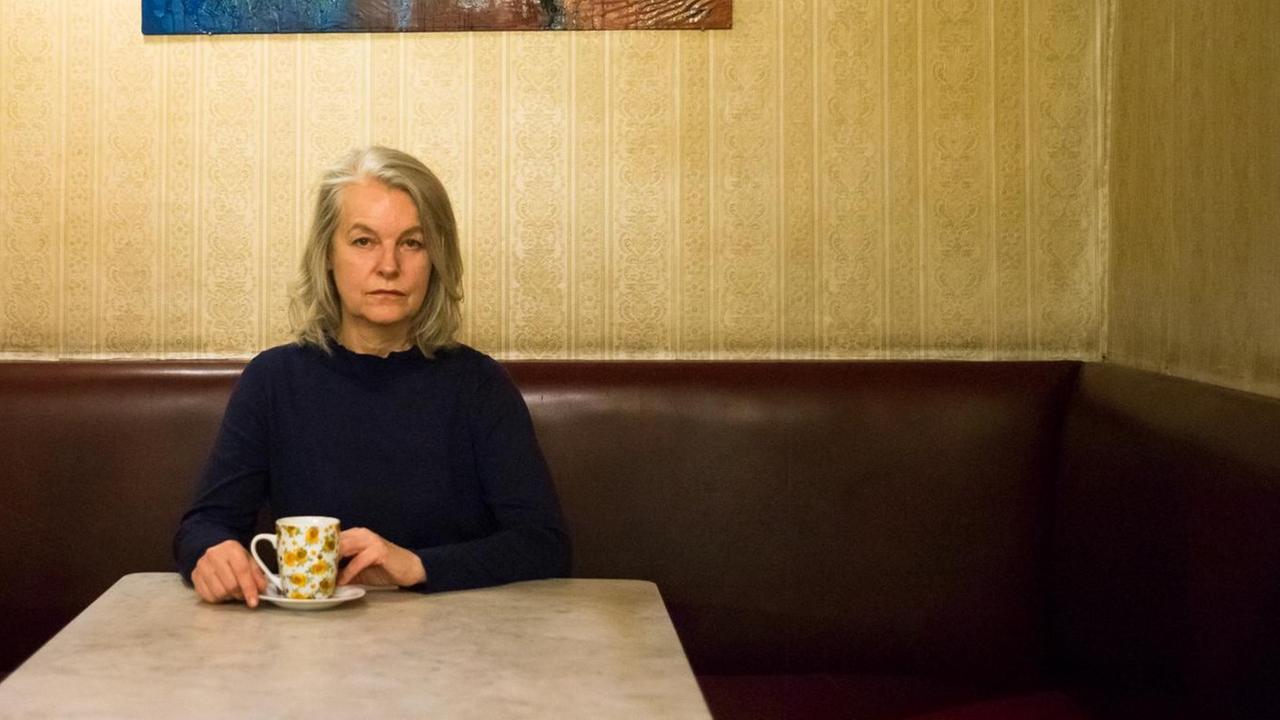 Eine Frau mit grauen Haaren sitzt in einem Café, hinter ihr an der Wand ist ein Gemälde. Sie hält in ihren Händen eine weiße Tasse.