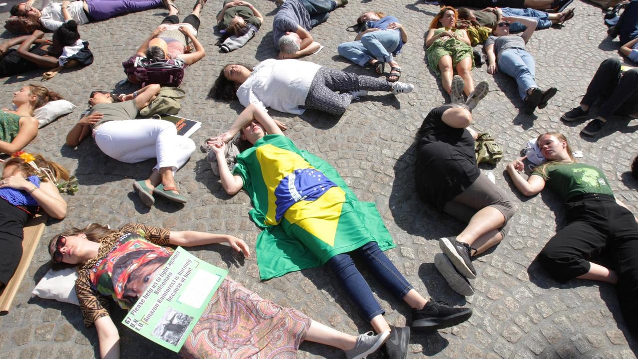 Menschen liegen während einer Demonstration gegen die Amazonas-Rodungen auf dem Boden, als ob sie gestorben seien. Eine Person ist in eine Brasilien-Flagge gehüllt, auf dem Schild einer anderen steht: Please help to fight against Bolsonaro. Amazon rainforest is burning because of him. Menschen liegen während einer Demonstration gegen die Amazonas-Rodungen auf dem Boden, als ob sie gestorben seien. Eine Person ist in eine Brasilien-Flagge gehüllt, auf dem Schild einer anderen steht: Please help to fight against Bolsonaro. Amazon rainforest is burning because of him.