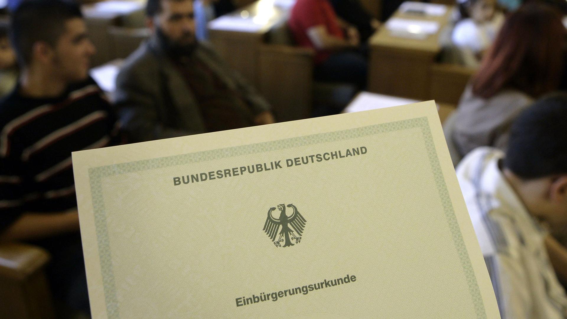 Die Einbürgerungsurkunde der Bundesrepublik Deutschland, im Hintergrund Migranten - aufgenommen bei einer Einbürgerungsfeier. Die Einbürgerungsurkunde der Bundesrepublik Deutschland, im Hintergrund Migranten - aufgenommen bei einer Einbürgerungsfeier.