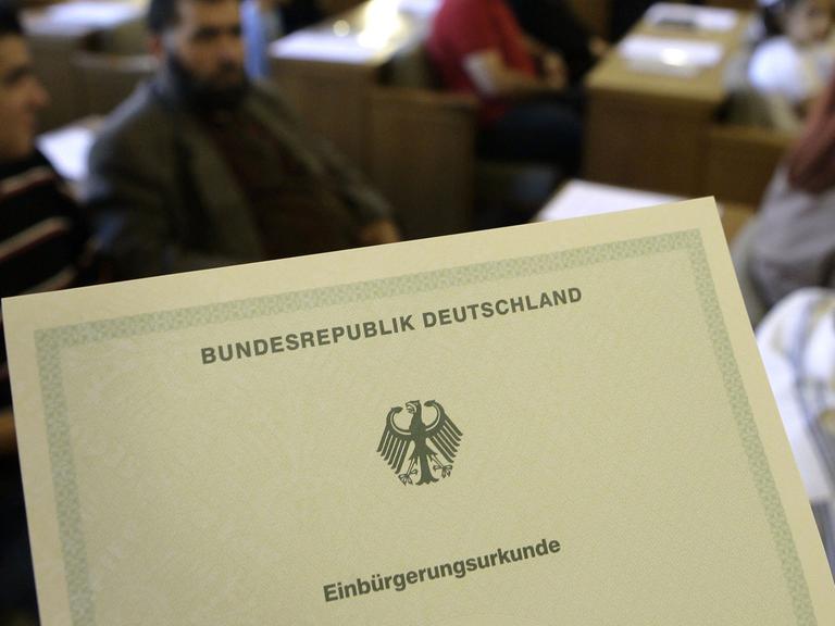 Die Einbürgerungsurkunde der Bundesrepublik Deutschland, im Hintergrund Migranten - aufgenommen bei einer Einbürgerungsfeier.