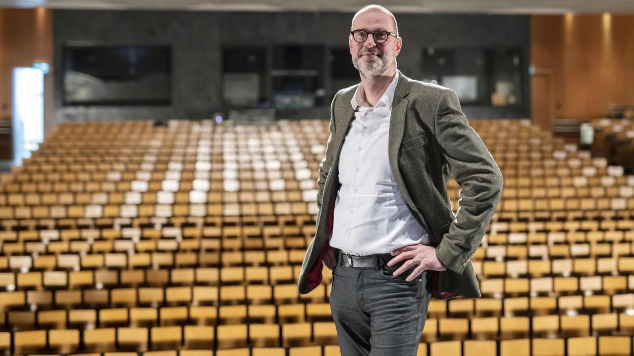 Ein Mann im Halbporträt mit Dreitagebart steht in einem Konzertsaal vor leeren, holzfarbenen Publikumsitzreihen. Ein Mann im Halbporträt mit Dreitagebart steht in einem Konzertsaal vor leeren, holzfarbenen Publikumsitzreihen.
