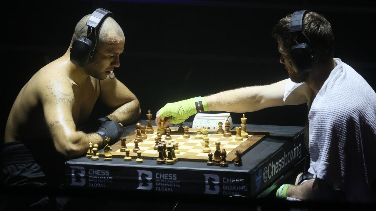 Zwei Männer sitzen an einem Schachspiel, sie haben Kopfhörer auf und sind verschwitzt; ihre Hände sind bandagiert. 