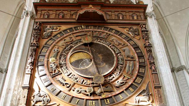 Eine alte astronomische Uhr von unten betrachtet in Großaufnahme. Zu sehen sind Holzschnitzereien, der Zeiger steht fast senkrecht.