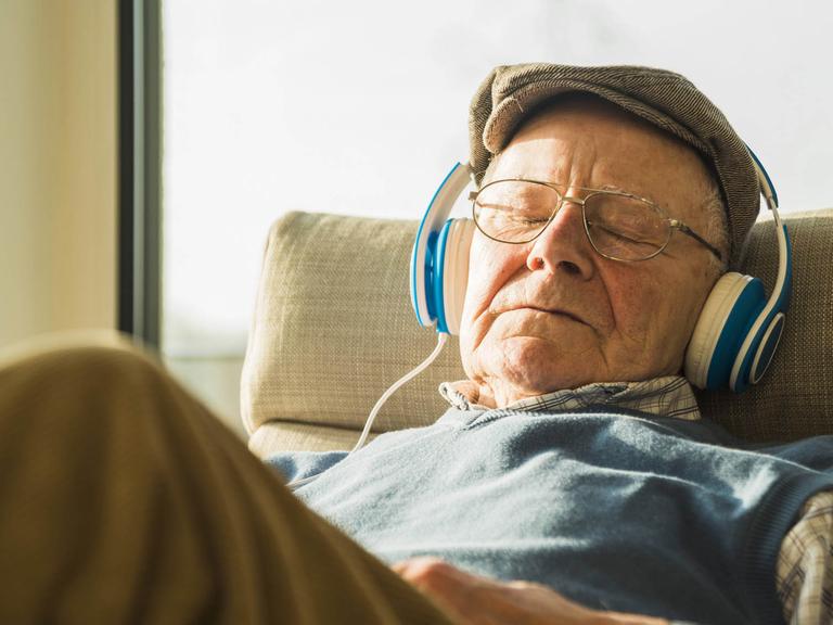 Ein Rentner sitzt mit Kopfhörern auf einem Sessel und hört Musik.
