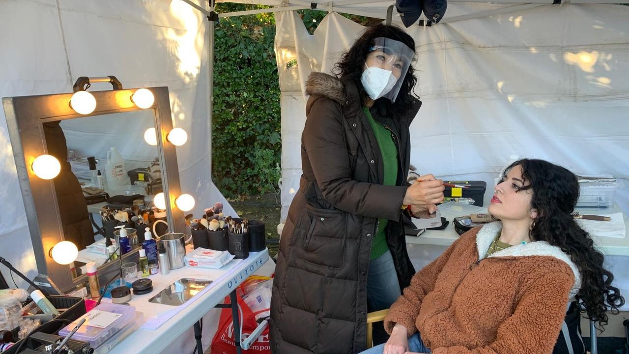 EIne Frau mit Maske und dickem Wintermantel schminkt eine Schauspielerin auf einem Stuhl.