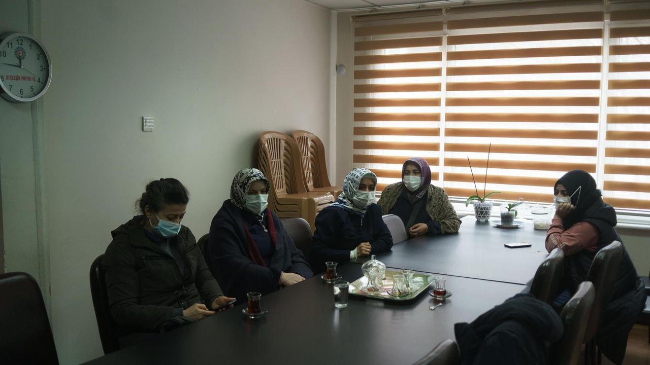 Mehrere Frauen sitzen mit Maske an einem langgezogenen dunklen Tisch. Mehrere Frauen sitzen mit Maske an einem langgezogenen dunklen Tisch.