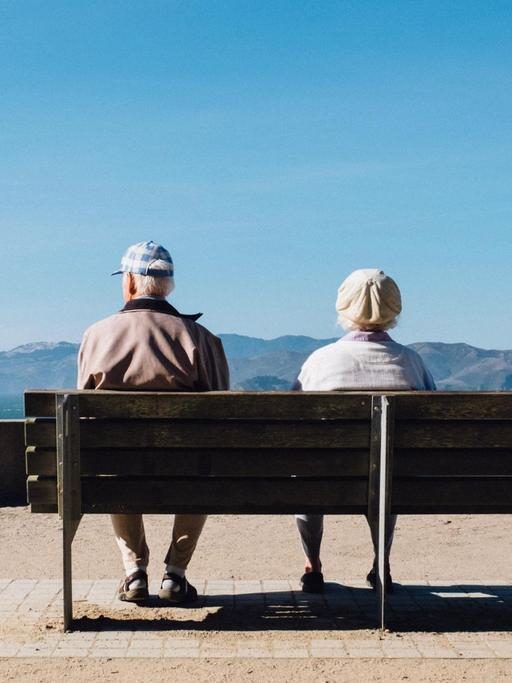 Zwei ältere Menschen sitzen auf einer Bank bei einer Aussichtsplattform und schauen auf die Berge.