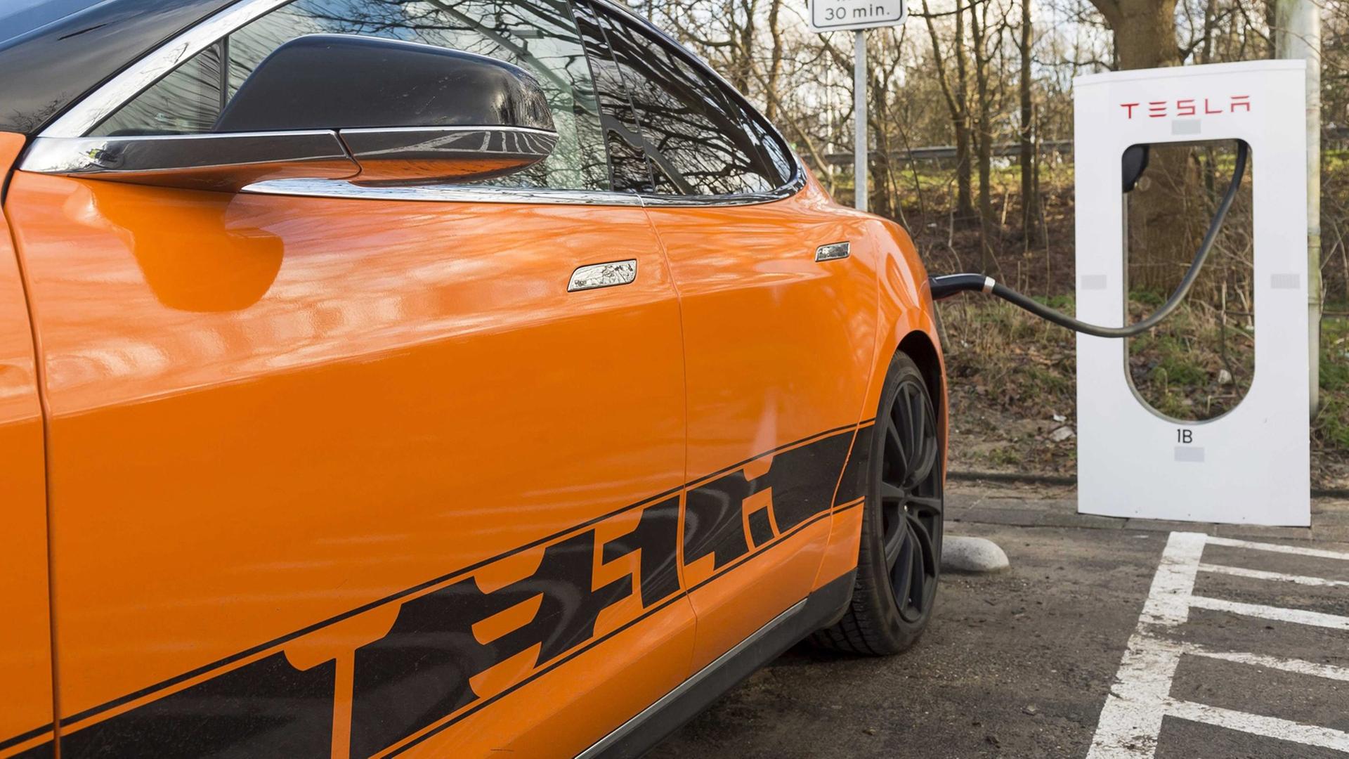 Ein orangefarbenes Elektro-Auto mit der Aufschrift "Tesla" ist an eine Lade-Station angeschlossen.