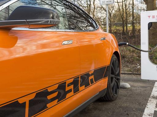 Ein orangefarbenes Elektro-Auto mit der Aufschrift "Tesla" ist an eine Ladestation angeschlossen.
