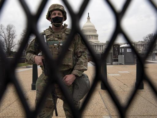 Durch einen Zaun hindurch ist ein Soldat zu sehen, der ein Gewehr vor sich hält. Im Hintergrund kann man das US-Kapitol in Washington mit seiner markanten weißen Kuppel sehen.