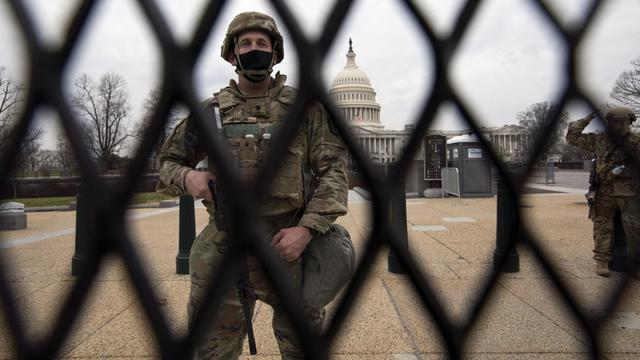 Durch einen Zaun hindurch ist ein Soldat zu sehen, der ein Gewehr vor sich hält. Im Hintergrund kann man das US-Kapitol in Washington mit seiner markanten weißen Kuppel sehen.