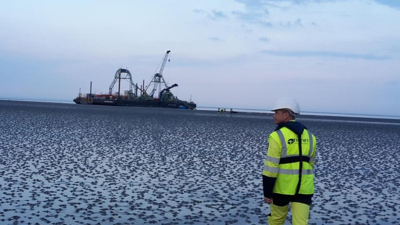 Ein imposantes Schiff bahnt sich seinen Weg durchs Wattenmeer, im Vordergrund läuft ein Arbeiter in typischer gelber Montur. Ein imposantes Schiff bahnt sich seinen Weg durchs Wattenmeer, im Vordergrund läuft ein Arbeiter in typischer gelber Montur.