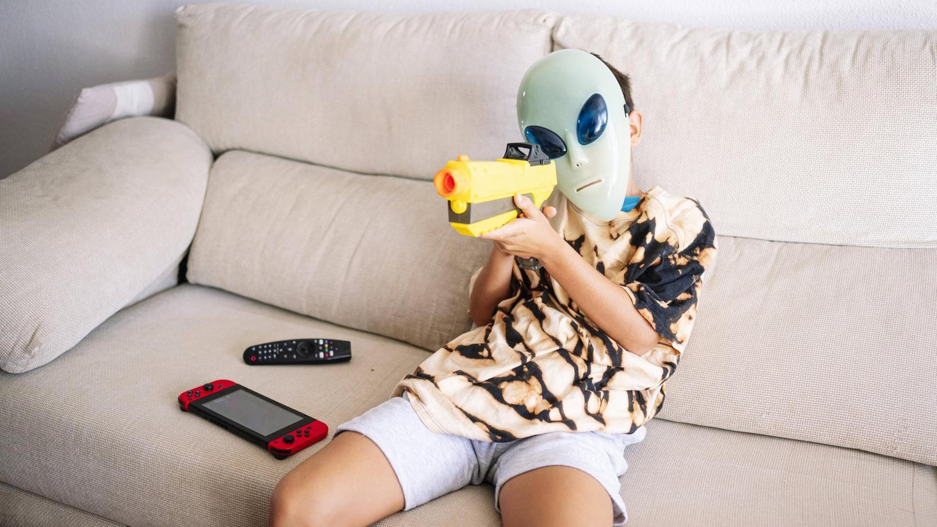 Ein Junge mit Alien-Maske und Plastikgewehr sitzt auf einem Sofa. file_source: imago / Westend 61 Ein Junge mit Alien-Maske und Plastikgewehr sitzt auf einem Sofa. file_source: imago / Westend 61