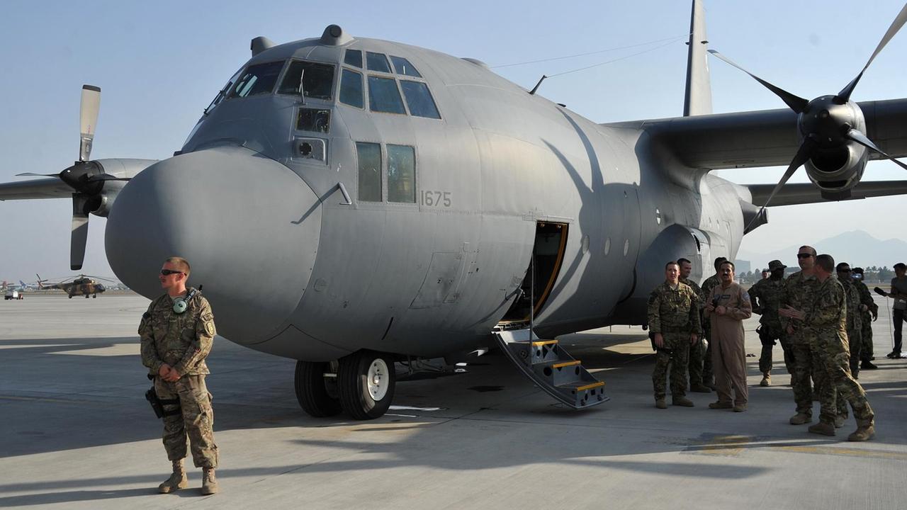 Vor der Maschine auf dem Rollfeld steht ein Soldat, an der Seite vor einer geöffneten Flugzeugklappe stehen weitere Soldaten.