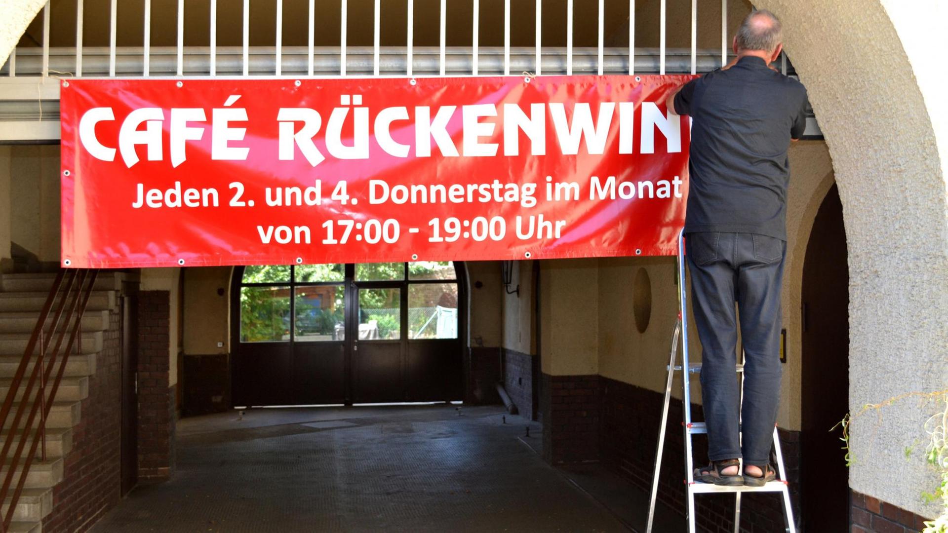 Stefan Friedrichowicz steht auf einer Leiter und hängt ein rotes Banner mit der Aufschrift "Cafe Rückenwind" auf. Stefan Friedrichowicz steht auf einer Leiter und hängt ein rotes Banner mit der Aufschrift "Cafe Rückenwind" auf.