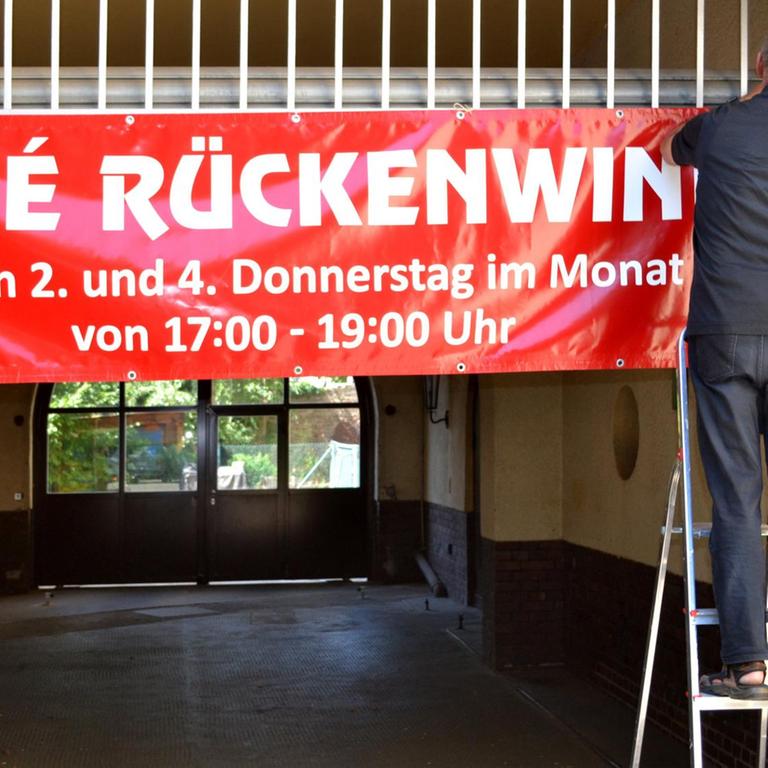 Stefan Friedrichowicz steht auf einer Leiter und hängt ein rotes Banner mit der Aufschrift "Cafe Rückenwind" auf.