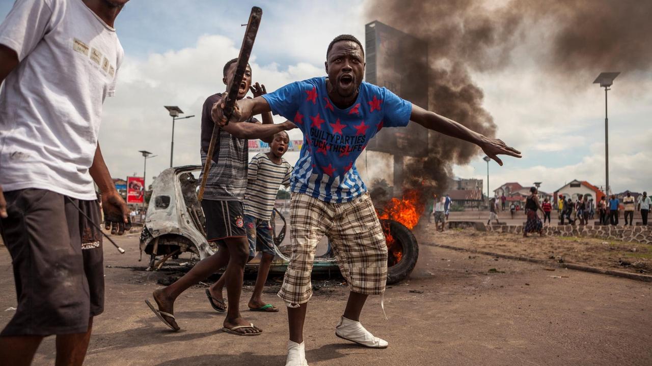 Ein Mann mit einem Stock bewaffnet, steht vor einem brennenden Fahrzeug in Kinshasa, der Hauptstadt der Demokratischen Republik Kongo.