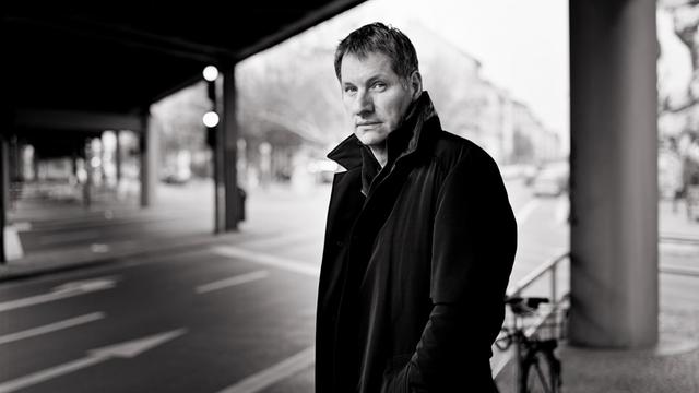 Marcus Wiebusch steht in einem dunklen Mantel unter einer Brücke an einer Strassenkreuzung.