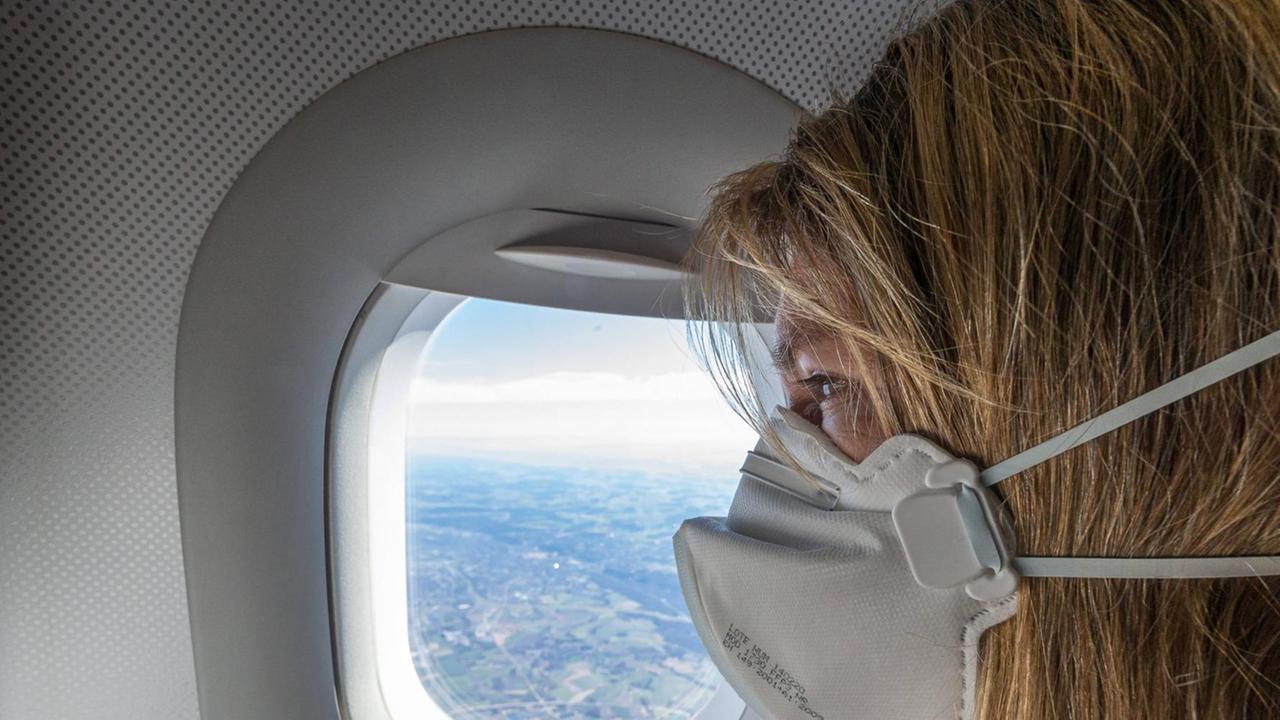 Eine Frau sitzt mit einer FFP2-Maske in einem Flugzeug am Fenster. Eine Frau sitzt mit einer FFP2-Maske in einem Flugzeug am Fenster.