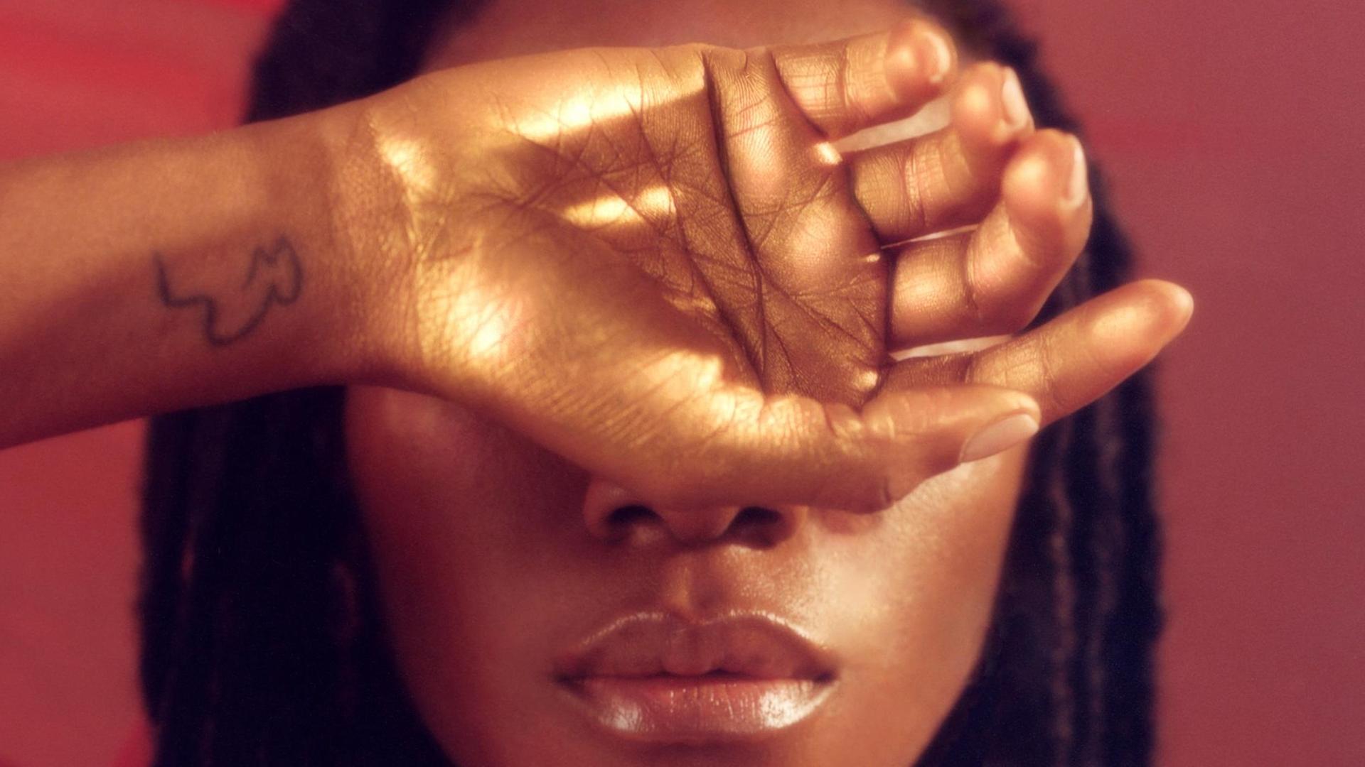Eine Frau hält sich eine Hand vor die Augen, die golden schimmert und am Handgelenk ein Tatoo zeigt. Eine Frau hält sich eine Hand vor die Augen, die golden schimmert und am Handgelenk ein Tatoo zeigt.