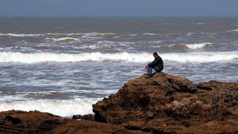 Ein Mann sitzt auf einem Felsen an der steinigen Atlantikküste von Essaouira.