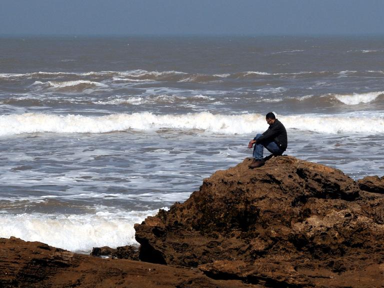 Ein Mann sitzt einsam auf einem Felsen und schaut aufs Meer.