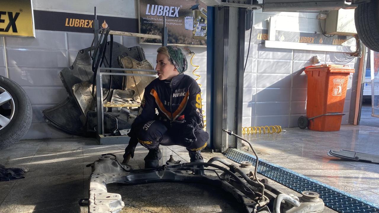 Eine junge Frau im Arbeitsoverall und mit Haarband hockt am Boden einer Autowerkstatt über einem Autoteil. Eine junge Frau im Arbeitsoverall und mit Haarband hockt am Boden einer Autowerkstatt über einem Autoteil.