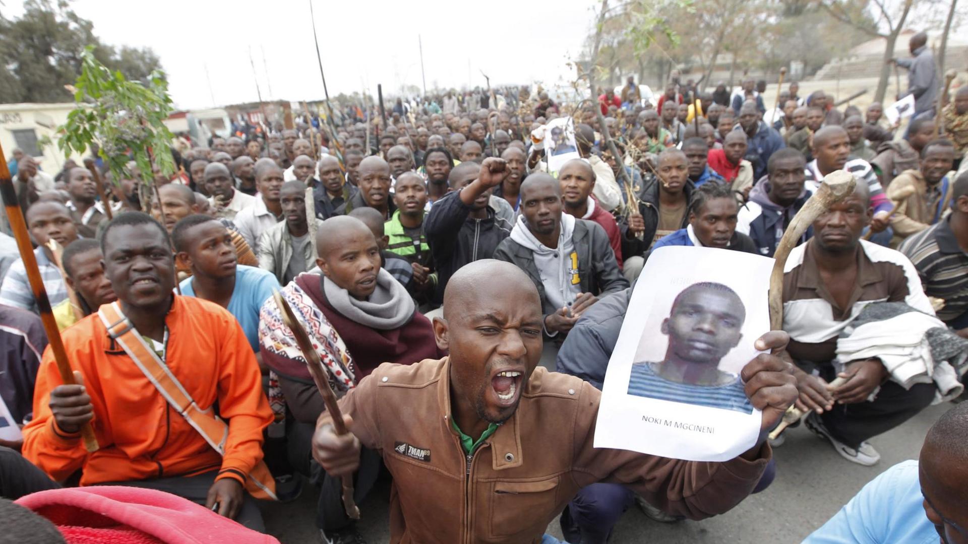 Eine große Menge afrikanischer Männer im Sitzstreik - im Vordergrund hält ein schreiender Mann das großformatige Foto eines jungen Mannes hoch, - es ist eines deerTodesopfer der blutigen Niederschlagung des Bergarbeiterstreiks südafrikanischen Marikanas