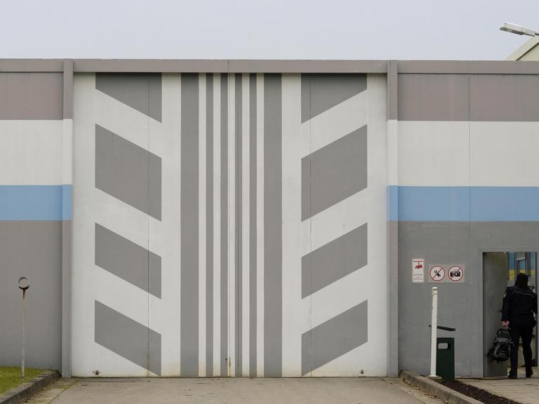 Ein Mann betritt die Jugendvollzugsanstalt Adelsheim in Baden-Württemberg. Das Rolltor hat ein grau-weißes Muster, die Mauer ist grau-weiß-blau gestrichen.