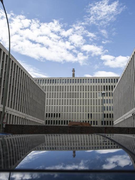 Der graue Neubau spiegelt sich in einem Autodach. Am blauen Himmel sind weiße Wolken.