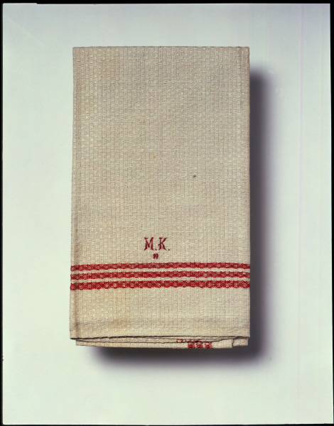 Weißes Leinenhandtuch mit roten Streifen im unteren Bereich und Monogramm in roten Buchstaben. Margarete Kuttner (1884-1944) gab dieses Handtuch ihrem Sohn Paul im Februar 1939 auf den Kindertransport nach England mit. Sie selbst wurde 1943 nach Auschwitz deportiert und ist dort umgekommen. Paul Kuttner bewahrte das Handtuch so gefaltet auf, wie er es bekommen hatte - als Erinnerungsstück an seine Mutter. Das Handtuch stammt aus der familieneigenen Textilfabrik S. Fränkel, Neustadt in Oberschlesien und gehörte vermutlich zur Aussteuer von Margarete Kuttner. Sie war eine geborene Fraenkel und mit der Familie der Textilunternehmer verwandt. Weißes Leinenhandtuch mit roten Streifen im unteren Bereich und Monogramm in roten Buchstaben. Margarete Kuttner (1884-1944) gab dieses Handtuch ihrem Sohn Paul im Februar 1939 auf den Kindertransport nach England mit. Sie selbst wurde 1943 nach Auschwitz deportiert und ist dort umgekommen. Paul Kuttner bewahrte das Handtuch so gefaltet auf, wie er es bekommen hatte - als Erinnerungsstück an seine Mutter. Das Handtuch stammt aus der familieneigenen Textilfabrik S. Fränkel, Neustadt in Oberschlesien und gehörte vermutlich zur Aussteuer von Margarete Kuttner. Sie war eine geborene Fraenkel und mit der Familie der Textilunternehmer verwandt.