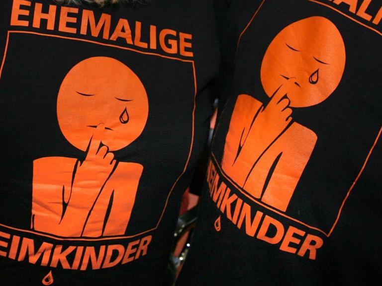 "Ehemalige Heimkinder" ist auf den T-Shirts eines Mannes und einer Frau zu lesen, auf denen auch eine weinende Figur aufgedruckt ist.