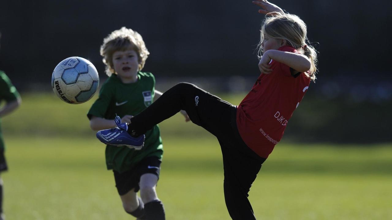 Fußballplatz: Im Vordergrund ein Mädchen mit einer artistischen Ballaktion. Im Hintergrund kommt ein kleiner Junge angesprintet. Fußballplatz: Im Vordergrund ein Mädchen mit einer artistischen Ballaktion. Im Hintergrund kommt ein kleiner Junge angesprintet.