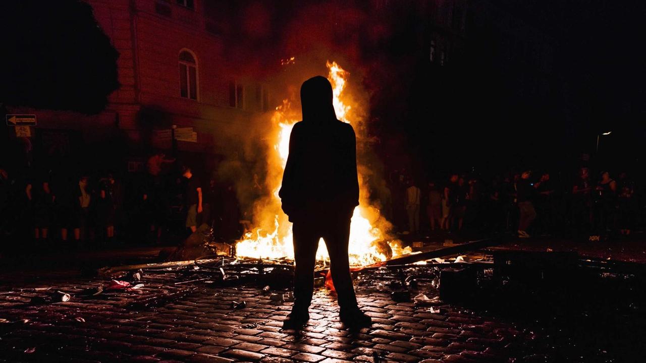 Ein Mann mit Kapuze steht nachts vor einem Feuer in einer Straße. Ein Mann mit Kapuze steht nachts vor einem Feuer in einer Straße.