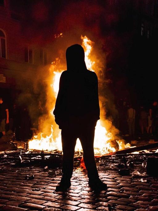Ein Mann mit Kapuze steht nachts vor einem Feuer in einer Straße.