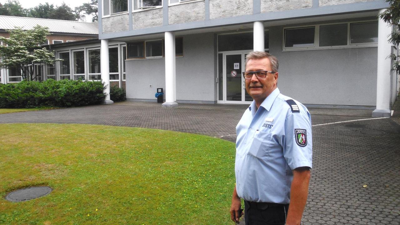 Frank Baucke steht in JVA Uniform auf einem gepflasterten Platz vor einem grauen Gebäude.