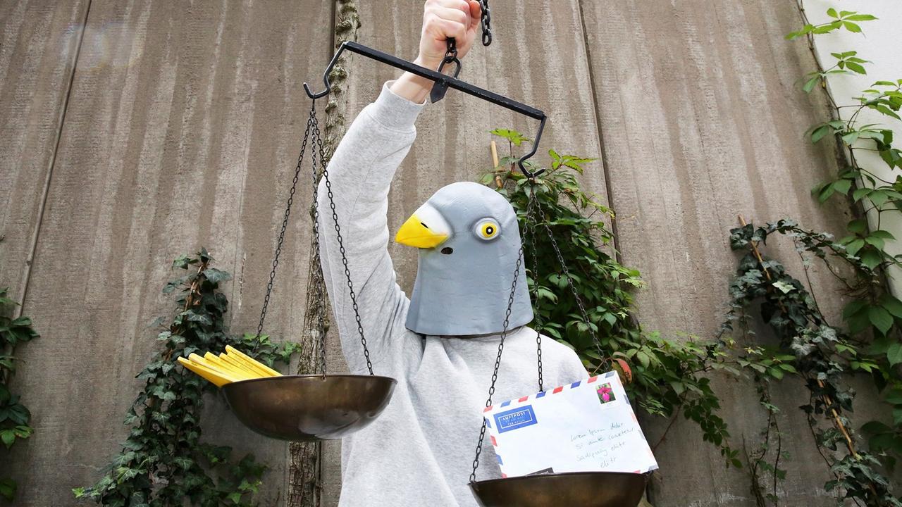 Ein Mensch trägt eine Maske, die einen Taubenkopf darstellt. In der rechten Hand hält die Taube eine Waage, in deren Schalen Briefe und gelbe Hefte liegen. Ein Mensch trägt eine Maske, die einen Taubenkopf darstellt. In der rechten Hand hält die Taube eine Waage, in deren Schalen Briefe und gelbe Hefte liegen.