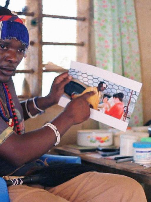 Ein Filmstill aus Afronautic Tales zeigt einen Afrikaner in einem blauen Monobloc sitzend, der auf eine Fotografie zeigt.