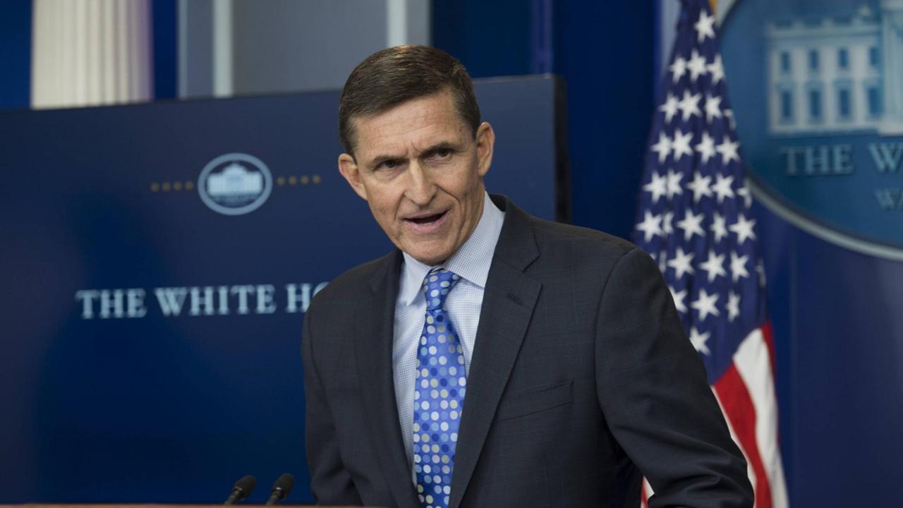 Flynn in einem Raum vor einer Tafel mit der Aufschrift "The White House" und einer US-.Flagge.
