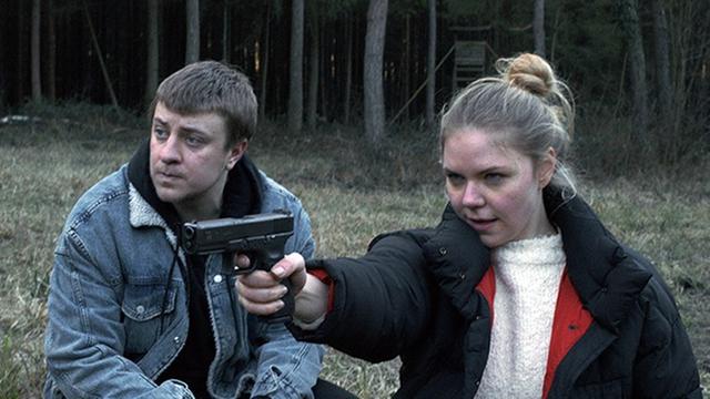 Eine Frau zielt im Wald mit einer Waffe. Ein Mann steht neben ihr.