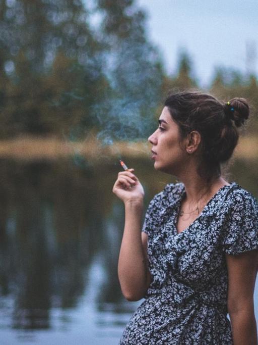 Eine junge Frau im Sommer steht an einem See und raucht.