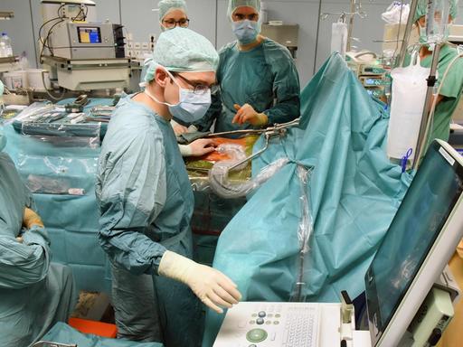 Eine Operation am Transplantationszentrum des Uniklinikums Leipzig. Medizinisches Personal in OP-Kleidung steht am OP-Tisch, eine Person bedient einen Computer.