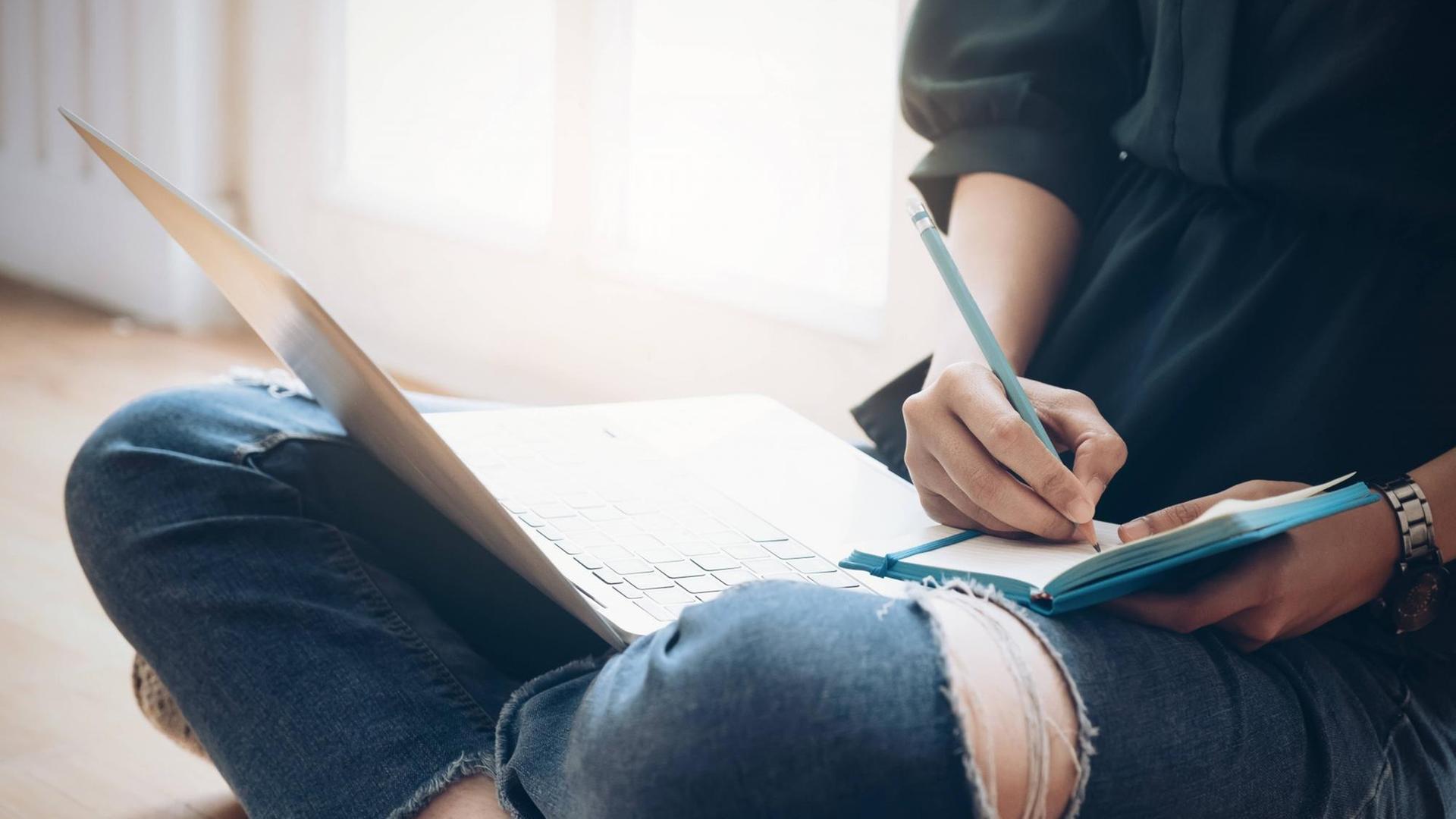 Eine Frau sitzt im Schneidersitz auf dem Boden, ein Notebook auf dem Schoß, und schreibt in ein Notitzbuch Eine Frau sitzt im Schneidersitz auf dem Boden, ein Notebook auf dem Schoß, und schreibt in ein Notitzbuch